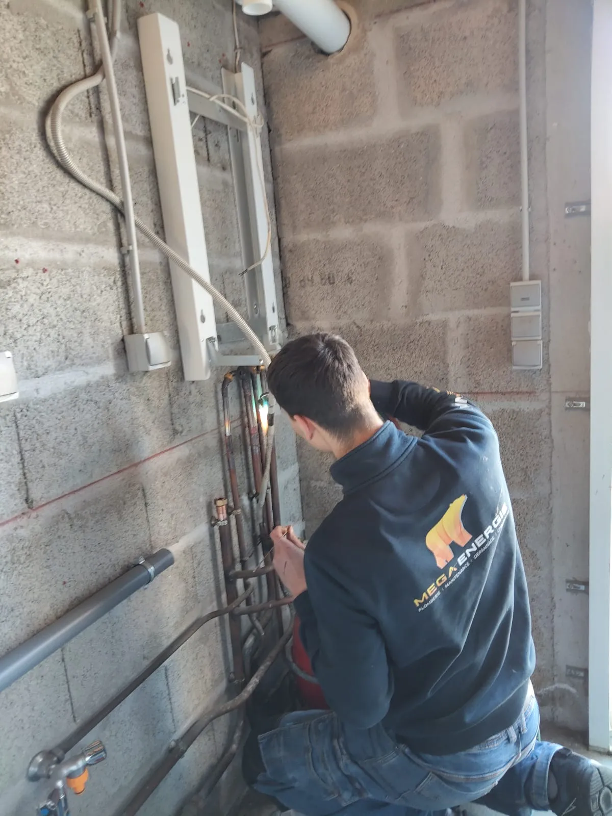 Un plombier chauffagiste de Mega Énergie de dos, réalisant un brasage au chalumeau sur des canalisations de cuivre fixées sur un mur en parpaings.
